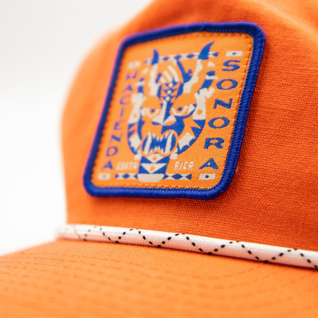 Orange baseball hat designed by Charlie Wagers depicting a traditional central american mask.