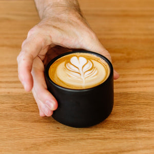 Matte black ceramic coffee mug from Fellow, held in a hand with latte art.