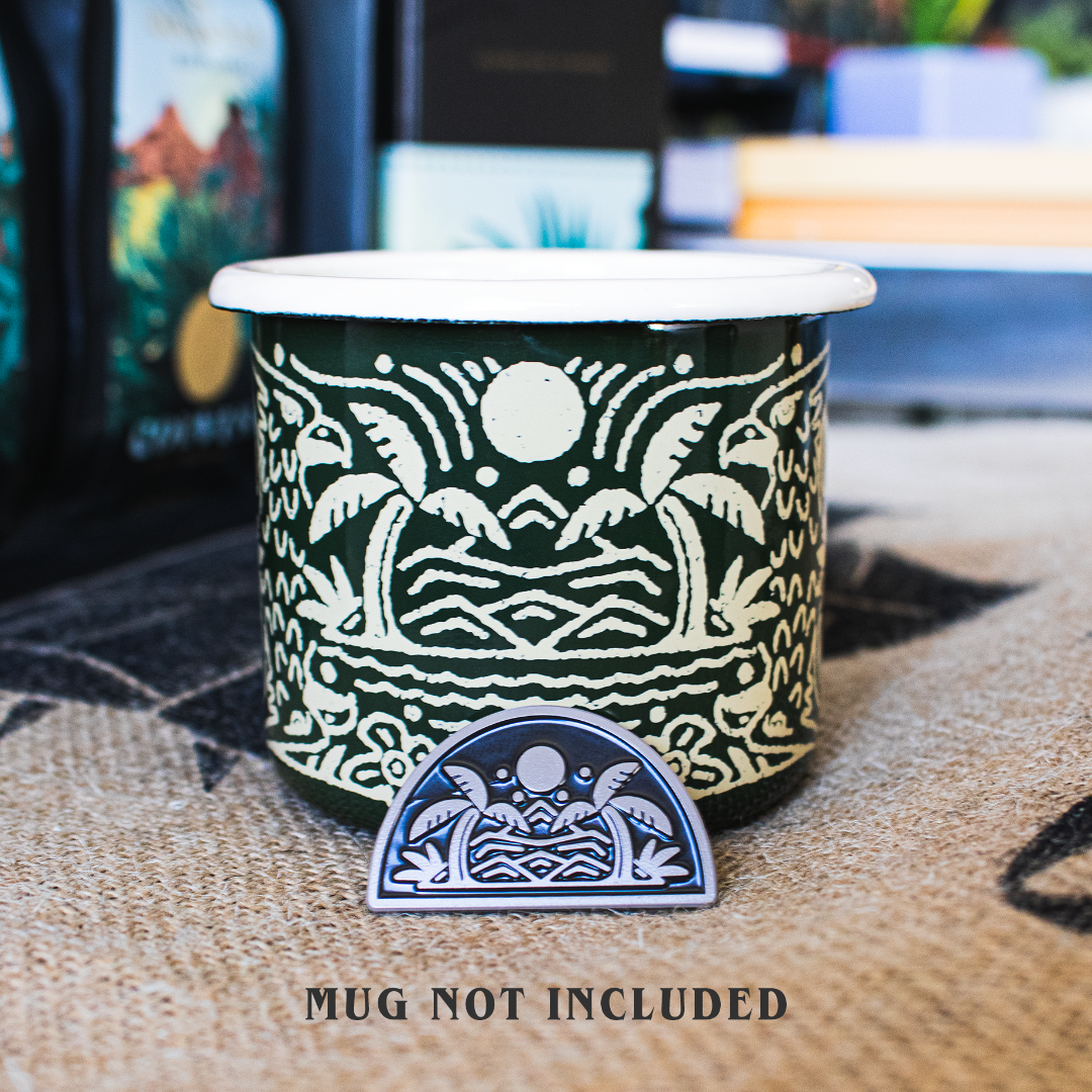 Enamel pin propped up against a metal coffee mug with coordinating designs of palm trees, waves, and mountains. 