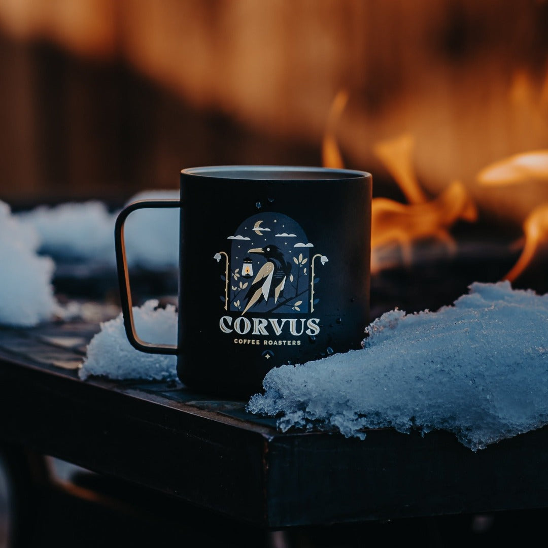 Specialty Coffee Roasters Camp Mug designed by Charlie Wagers depicting a crow in a night scene holding a lantern, sitting in the snow by a firepit.