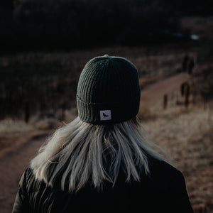 Woman outside wearing a green wool beanie with a crow logo.