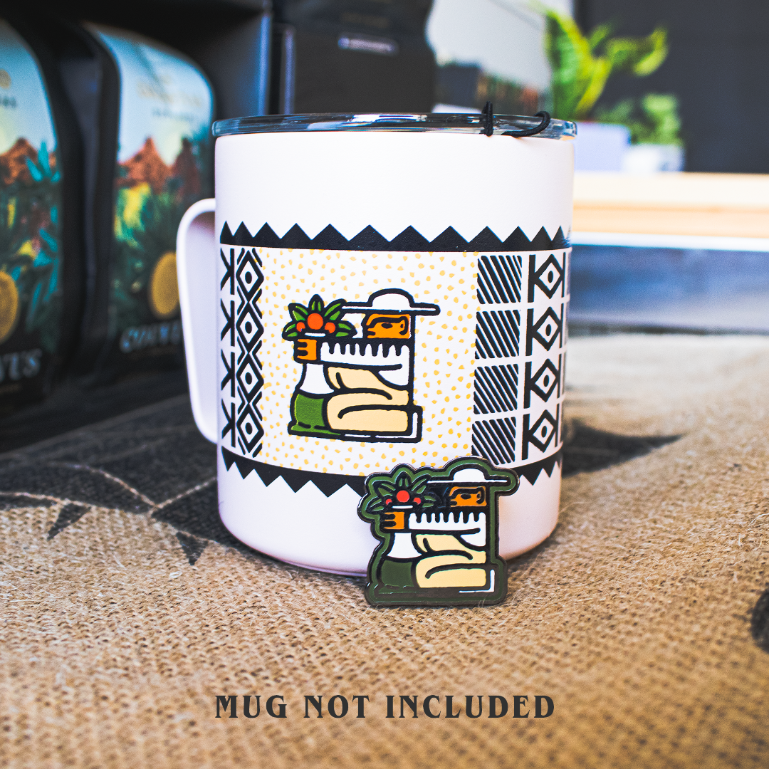 Enamel pin propped up against a metal coffee mug with coordinating designs of a coffee farmer..