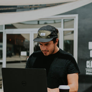 Man wearing a five-panel hat while sitting at a computer in front of a coffee shop.